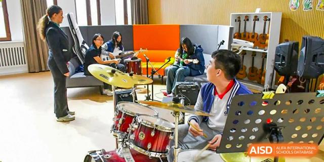 Students at Oriental Cambridge International School playing instruments, including a drum set, in an indoor music practice room during band rehearsal.