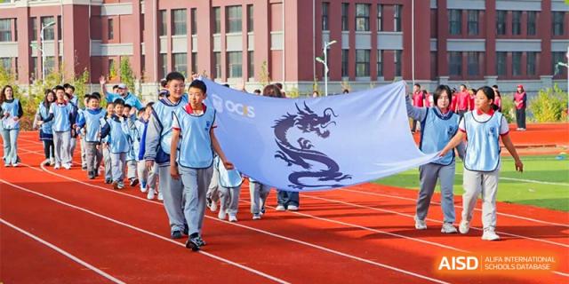 Students from Oriental Cambridge International School marching on a red running track while holding a large white banner with the school's dragon logo on it.