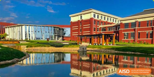 An exterior view of a large red and brick school building at Oriental Cambridge International School, with its reflection visible in the adjacent pond under a clear blue sky.