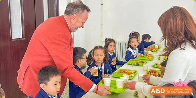 A teacher or adult at Oriental Cambridge International School helping a small group of young students with their meals and drinks in the school cafeteria or dining hall.
