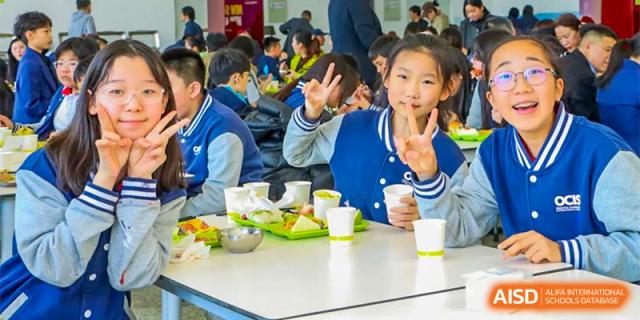 A female student in a blue and white uniform at Oriental Cambridge International School smiling and making a peace sign while seated at a dining table with her meal.