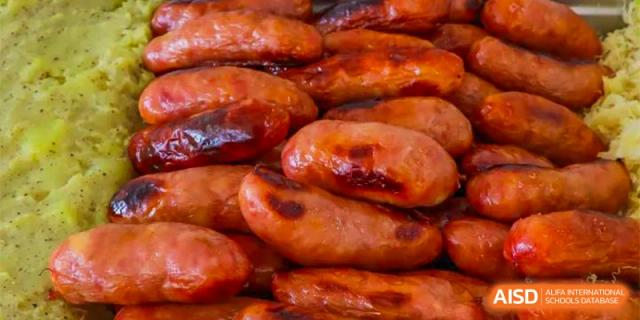 A close-up of a tray filled with glazed, reddish-brown sausages or hot dogs, a food option available in the cafeteria at Oriental Cambridge International School.