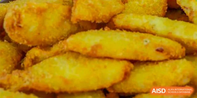 A close-up of a serving tray of golden-brown baked or fried potato wedges or similar food in the cafeteria at Oriental Cambridge International School.