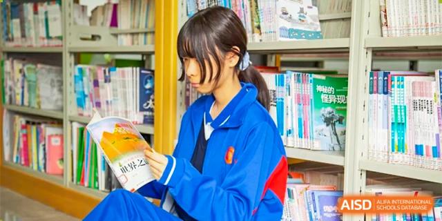 A female student with a ponytail wearing a blue athletic jacket sits in a library and carefully reads through a textbook at Guangzhou Yinghao School.