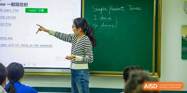 A teacher in a striped shirt points to a chalkboard while explaining the simple present tense during an English class at Guangzhou Yinghao School.