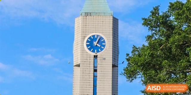 A tall, modern white clock tower with a blue circular face stands prominently against a clear blue sky on the campus of Guangzhou Yinghao School.