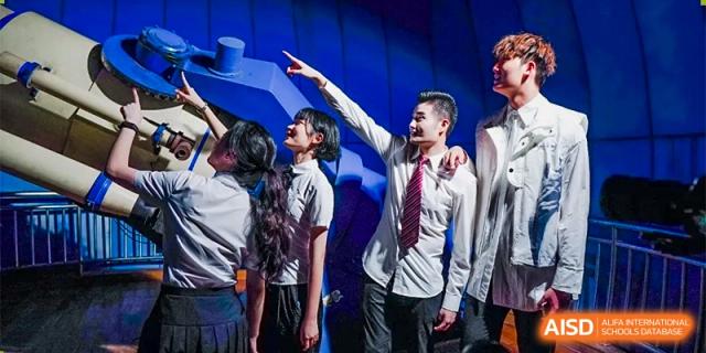 Students in formal school uniforms stand on a stage under blue lighting, gesturing upward during a performance at Guangzhou Yinghao School.