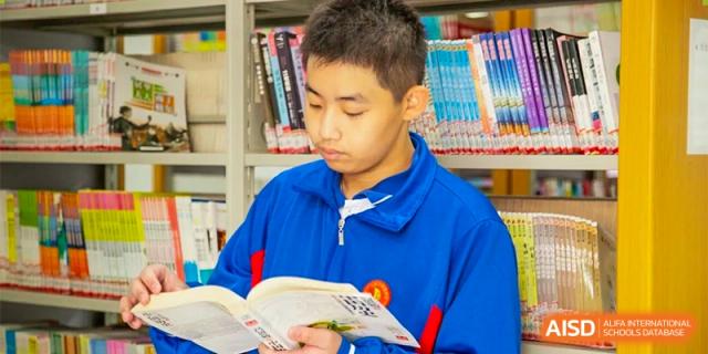 A male student in a blue track jacket stands in front of bookshelves, concentrating on the pages of a book he is holding at Guangzhou Yinghao School.