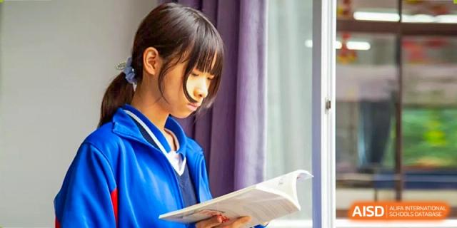 A close-up shot of a female student in a blue jacket looking down with focus while studying a textbook indoors at Guangzhou Yinghao School.