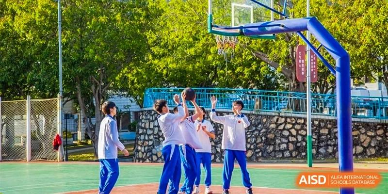 Students in white and blue uniforms play basketball on an outdoor court surrounded by lush green trees during a physical education class at Guangzhou Yinghao School.