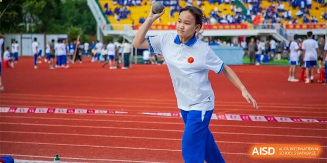 A female student in a white polo and blue athletic pants prepares to throw a shot put during a track and field sports competition at Guangzhou Yinghao School.