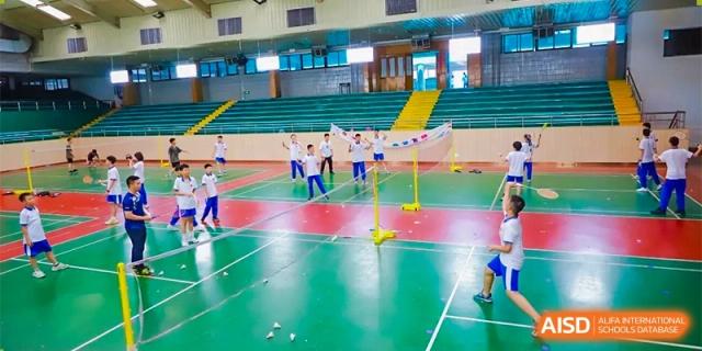Students play badminton on multiple green indoor courts under bright gymnasium lighting at Guangzhou Yinghao School.