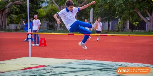 A student in a white and blue athletic uniform successfully clears a high jump bar during a track and field event at Guangzhou Yinghao School.