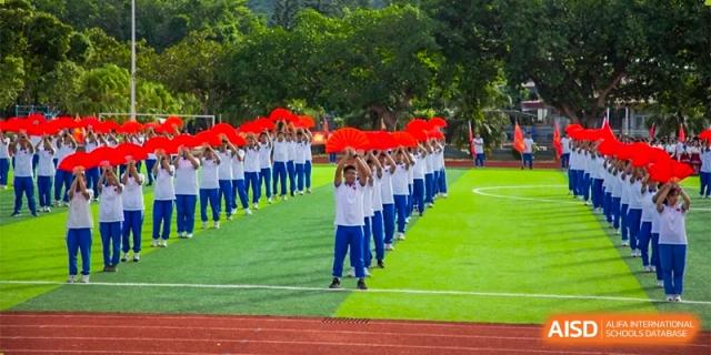 A large line of students in white and blue uniforms perform a synchronized routine with red fans on a green field at Guangzhou Yinghao School.