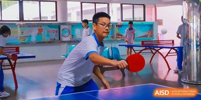 A student wearing glasses focuses intently while playing a competitive game of table tennis in an indoor facility at Guangzhou Yinghao School.