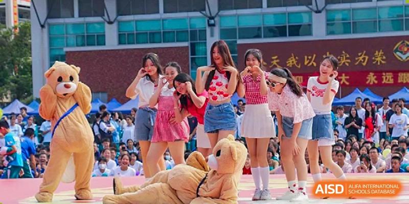 A group of female students performing on stage outdoors, with one wearing a teddy bear costume, at Guangdong Experimental High School IFPAD & A-Level.