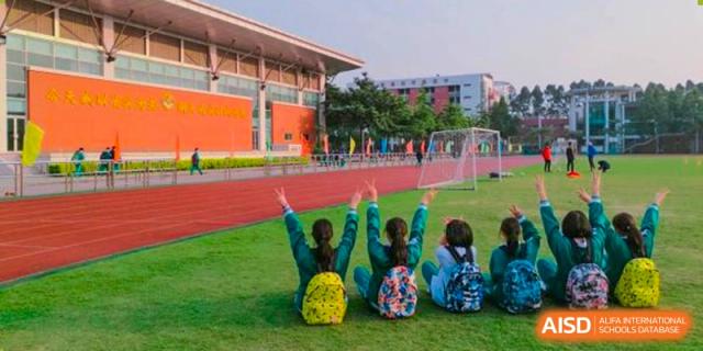 Three students with backpacks sitting on the grass of a sports field, cheering with their arms raised, with school buildings in the background at Guangdong Experimental High School IFPAD & A-Level.
