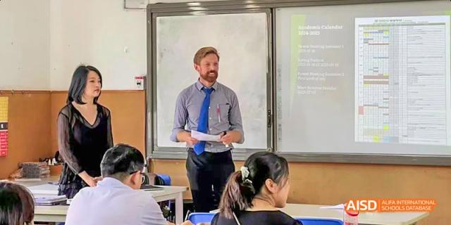 A male teacher in a dress shirt and tie teaching a class, standing in front of a whiteboard while addressing students, at Beanstalk International Bilingual School.
