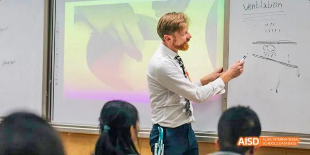 A male teacher with a beard standing next to a whiteboard, gesturing with his hand while teaching a lesson to students, at Beanstalk International Bilingual School.
