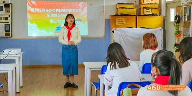 A female student wearing a white shirt and red scarf standing up and speaking or presenting in a brightly lit classroom to her seated classmates, at Beanstalk International Bilingual School.