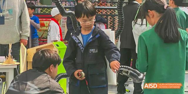 A male student in a dark jacket with a "FILA" logo is focused on a hands-on technical or science project indoors, at Beanstalk International Bilingual School.