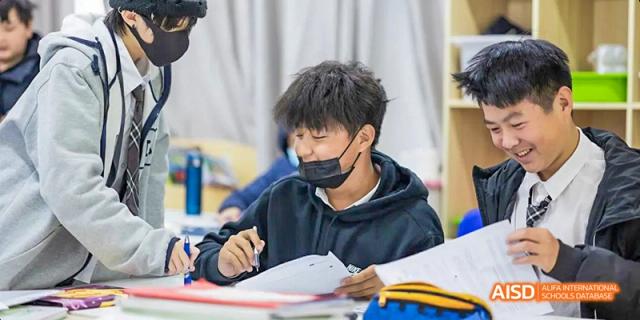 A male student wearing a black hoodie and a face mask is focused on writing notes or completing an assignment in a classroom setting, at Beanstalk International Bilingual School.