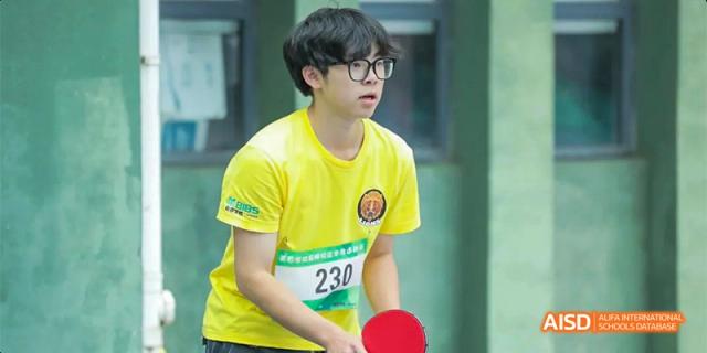 A young male student wearing a yellow shirt and glasses, holding a ping pong paddle and focused on the game, at Beanstalk International Bilingual School.