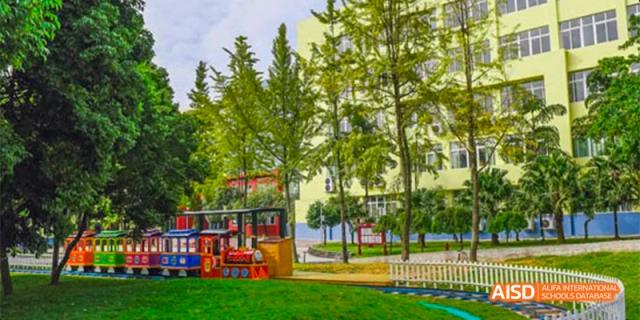 A vibrant outdoor playground with green grass and tall trees, next to a yellow and blue school building, at Beanstalk International Bilingual School.