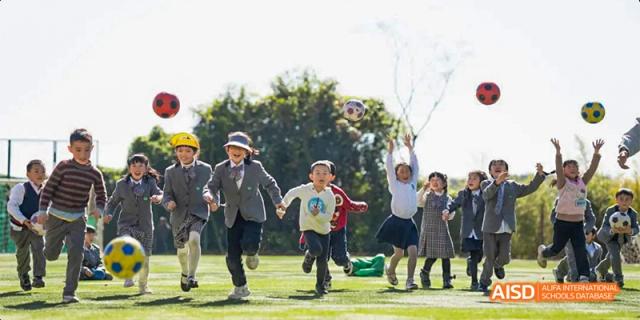A group of young students running and playing happily on a sunny green field, with one girl throwing a ball in the air, at Beanstalk International Bilingual School.
