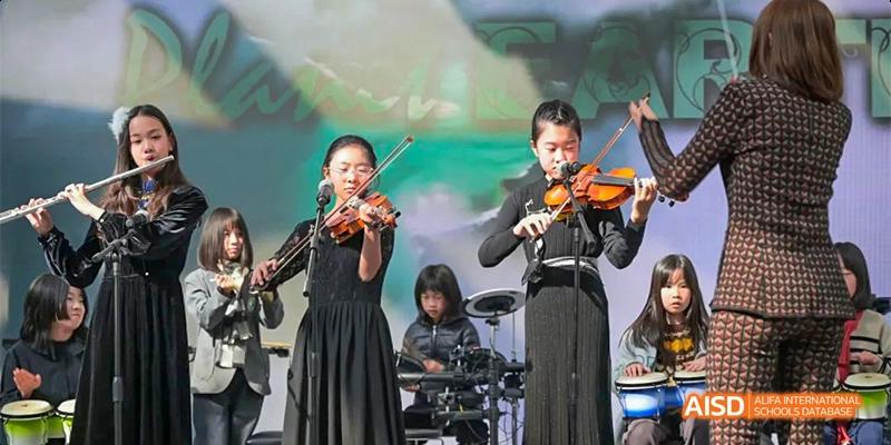 Three musicians, two adults and one child, dressed in black performing with violins and a drum kit on an outdoor stage, at Beanstalk International Bilingual School.