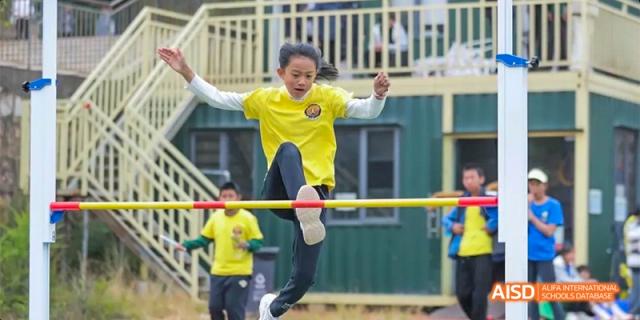A young girl in a yellow shirt and black shorts performing a high jump or a similar athletic maneuver over a bar, at Beanstalk International Bilingual School.
