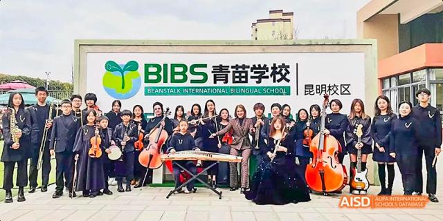 A large group of students and adults posing together with musical instruments outdoors, with a banner behind them that says "BIBS 菁苗学校," at Beanstalk International Bilingual School.