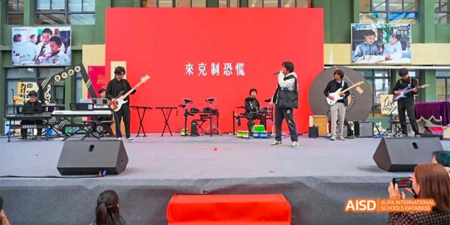 A student performing or speaking on a stage with a bright red backdrop and a sign in Chinese characters, with musicians sitting in the background, at Beanstalk International Bilingual School.