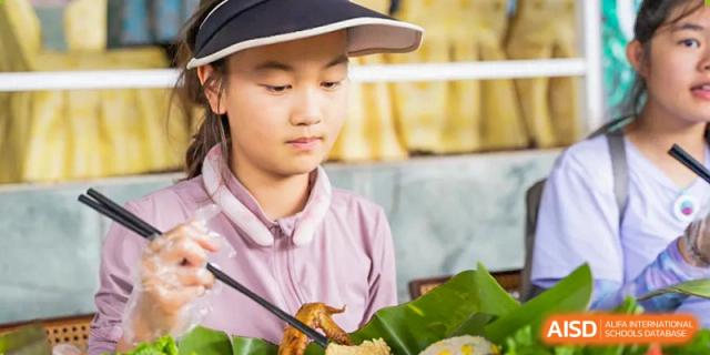A female student wearing a visor hat is carefully eating a meal with chopsticks, focusing on the food on her plate, at Beanstalk International Bilingual School.