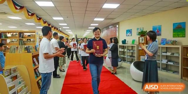 A smiling woman walks down a red carpet in a hallway, holding a certificate as others applaud her achievement at Hefei High-tech Sino-Canadian School.