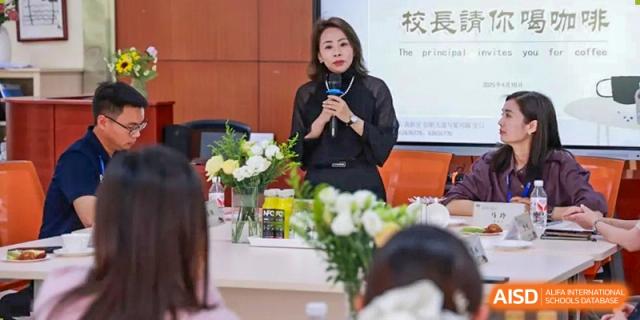 A woman in professional attire speaks into a microphone at a formal gathering, with flowers and refreshments on the table at Hefei High-tech Sino-Canadian School.