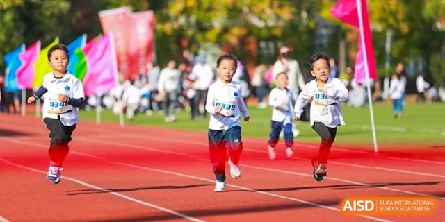 Young children in white jerseys race down the red running track during an outdoor athletic competition at Hefei High-tech Sino-Canadian School.