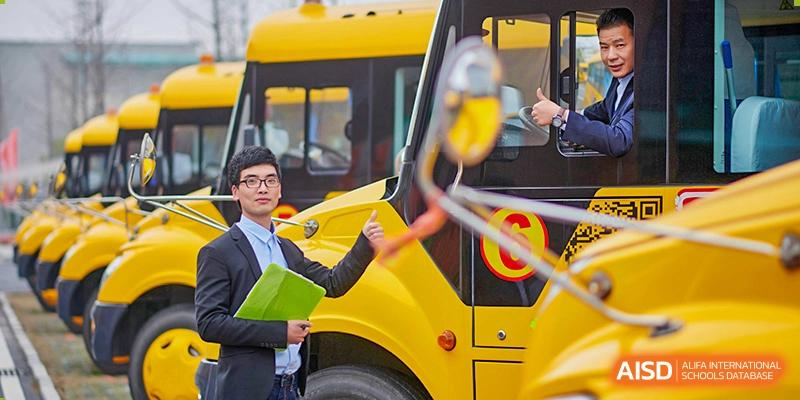 A teacher in a suit stands next to a bright yellow school bus, giving a thumbs up to signify safety at Hefei High-tech Sino-Canadian School.