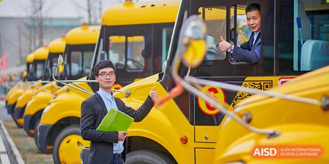 A teacher in a suit stands next to a bright yellow school bus, giving a thumbs up to signify safety at Hefei High-tech Sino-Canadian School.