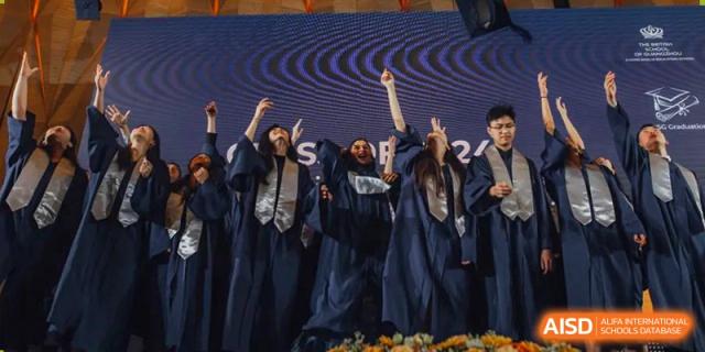 Graduating students from The British School of Guangzhou in navy blue gowns cheering with their arms raised on a stage.