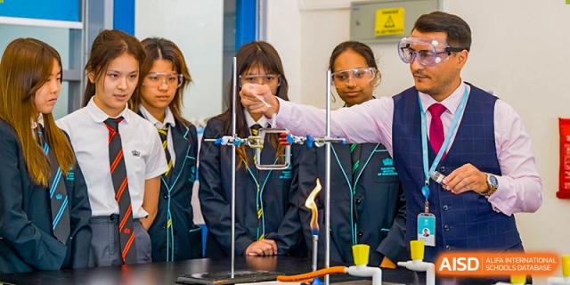 Students and a teacher at The British School of Guangzhou wearing safety goggles while conducting a chemistry experiment involving an open flame.