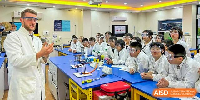 A classroom of The British School of Guangzhou students in lab coats and safety goggles, attentively watching a science experiment demonstration.