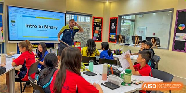Young students and a teacher at The British School of Guangzhou engaged in a lively activity in a bright classroom, with a teacher demonstrating a movement.
