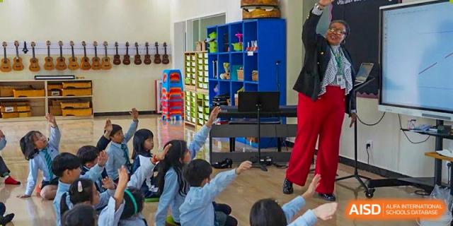 Younger students from The British School of Guangzhou gathered in a brightly colored music or activity room, raising their hands.
