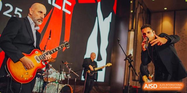 A musician playing an electric guitar on a stage with bright red and black graphics in the background, likely at a school event for The British School of Guangzhou.