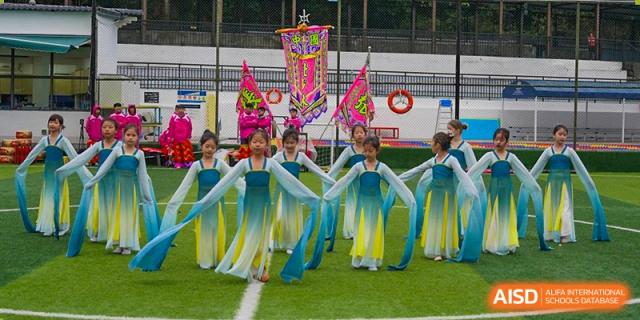 Students from The British School of Guangzhou performing a traditional fan or ribbon dance in colorful costumes on an outdoor field.