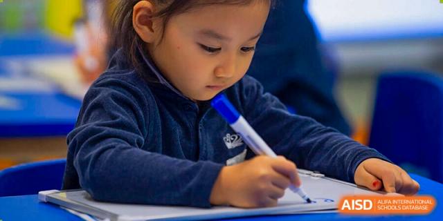A young child from The British School of Nanjing in a dark blue shirt, writing on paper with a blue marker.