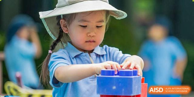 A young child from The British School of Nanjing in a blue shirt and white sun hat playing with large blue and red blocks.