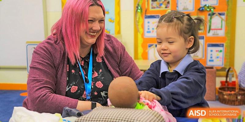 A female teacher from The British School of Nanjing with pink hair, holding a doll while teaching a young child.
