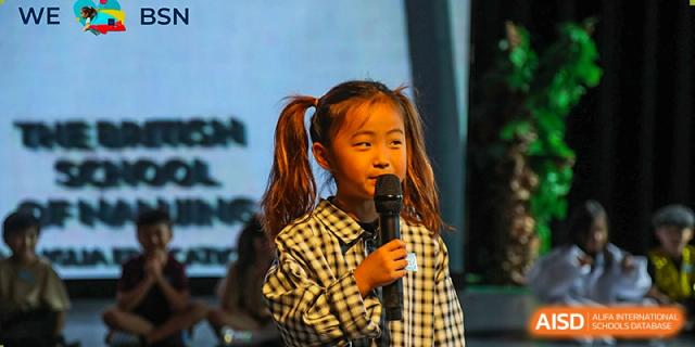 A young student from The British School of Nanjing with pigtails, holding a microphone and singing on a stage.
